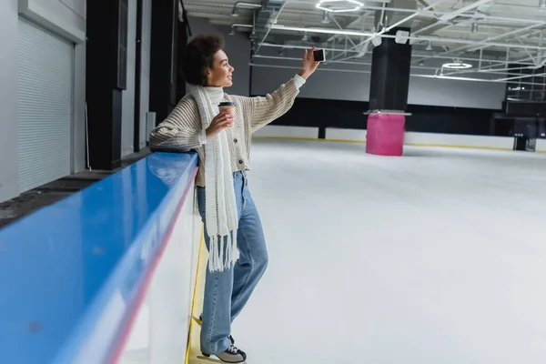 アイススケートにおけるアフリカ系アメリカ人女性の側面図紙コップを保持し アイスリンク上のスマートフォンで自撮り — ストック写真