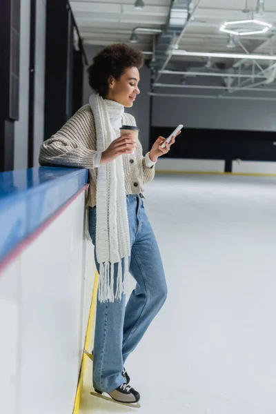 Side view of smiling african american woman in ice skates using mobile phone and holding coffee to go on ice rink