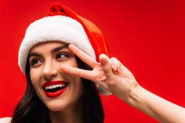 Noel Baba şapkalı neşeli genç kadın kırmızıda izole edilmiş zafer işareti gösteriyor.