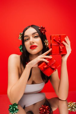 Young model in top and gift bows on hair holding presents near reflective surface isolated on red  clipart