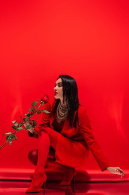 Kırmızı arka planda gül tutan topuklu ve inci kolyeli şık bir kadın. 