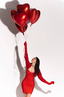 Cheerful woman in dress and gloves looking at red balloons in heart shape on white background with shadow  clipart