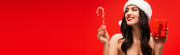 Красивая модель в шляпе Санта держа подарок и полосатый леденец изолирован на красный, баннер 