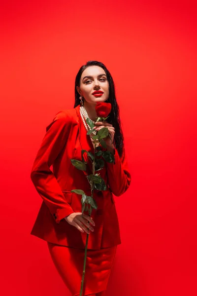 Stylish woman in pearl necklace holding rose on red background