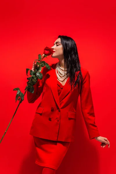 Young woman in jacket and pearl necklace smelling rose on red background