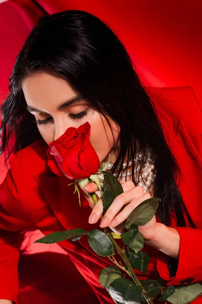 Brunette woman in pearl necklace holding rose near face on red background