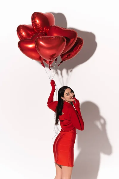 Cheerful brunette model in red dress and gloves looking at camera near heart shaped balloons on white background with shadow