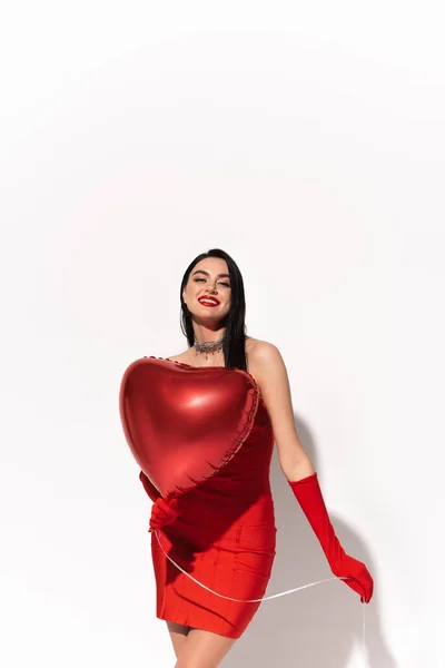 Pretty brunette woman in red gloves and dress holding heart shaped balloon on white background with shadow