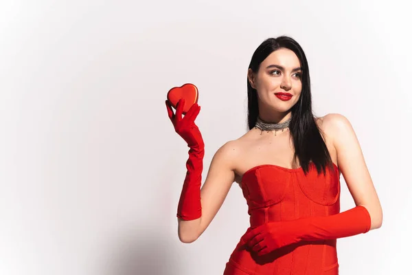 Stylish woman in red gloves and dress holding heart shaped present on grey background