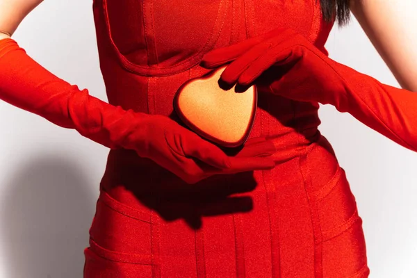 Cropped view of woman in red gloves and dress holding present in heart shape on grey background