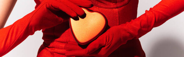 Partial view of woman in red gloves holding heart shaped gift box on grey background, banner 