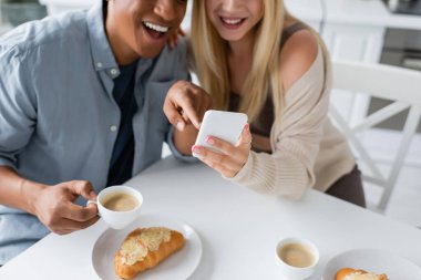 partial view of laughing african american man pointing at smartphone near coffee cups and tasty croissants clipart