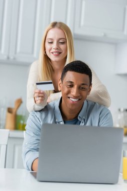 Mutfakta, gülümseyen Afrikalı Amerikalı erkek arkadaşı ve dizüstü bilgisayarın yanında kredi kartı tutan sarışın bir kadın.