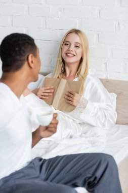 Kitabı olan genç ve neşeli bir kadın bulanık Afrikalı Amerikalı bir adama bakıyor. Elinde kahve bardağıyla yatak odasında.