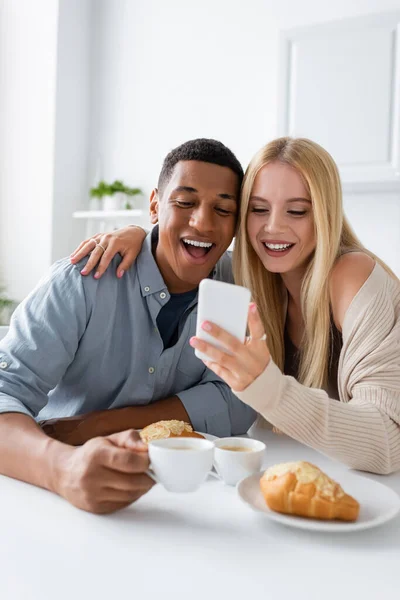 兴奋的跨种族夫妇在早餐时一边看手机一边笑 — 图库照片