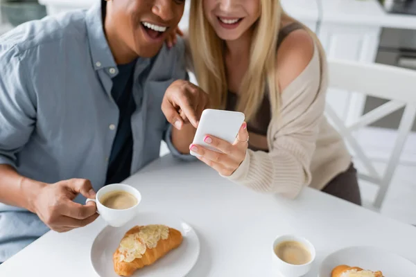 stock image partial view of laughing african american man pointing at smartphone near coffee cups and tasty croissants