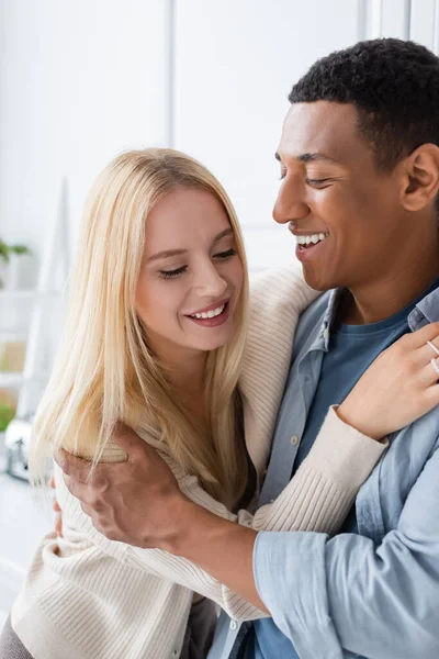 stock image cheerful blonde woman and smiling african american man embracing in kitchen
