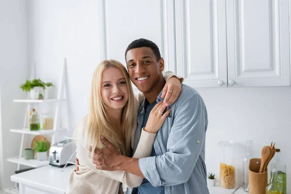 在厨房拥抱时看着相机的快乐的跨种族夫妇 — 图库照片