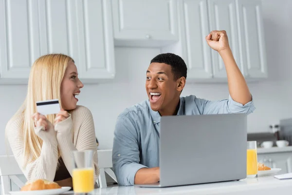 興奮したアフリカ系アメリカ人の男が喜んでノートパソコンとガールフレンドとクレジットカードで朝食をキッチンで — ストック写真