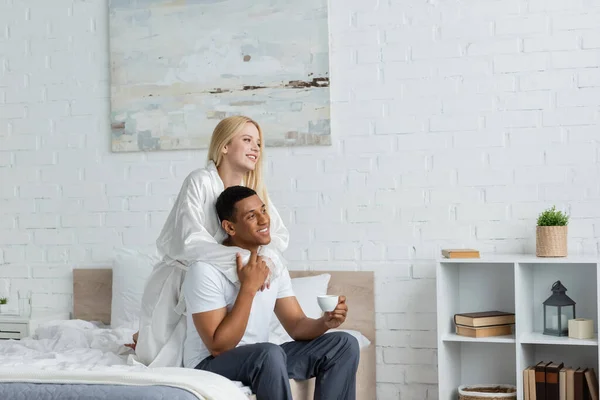 young woman in white silk robe hugging african american boyfriend sitting on bed with morning coffee