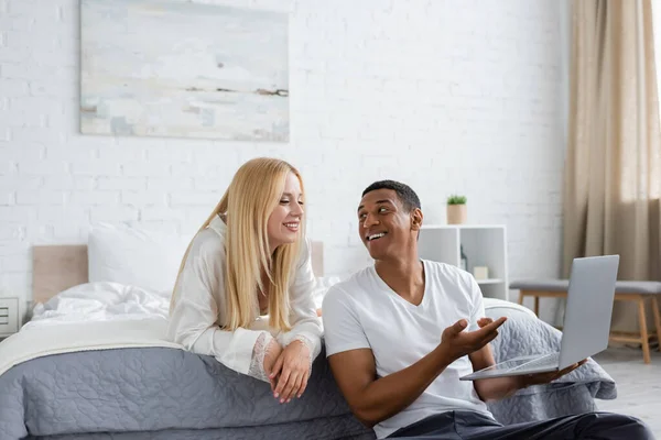 陽気なアフリカ系アメリカ人男性がノートパソコンを指差してベッドルームで笑顔の彼女を見て — ストック写真