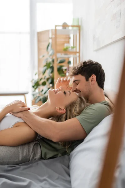 Side view of cheerful man in pajama hugging blonde girlfriend on bed in morning