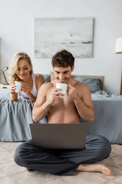 積極的な筋肉男保持カップのコーヒーと見ますラップトップ近くのガールフレンドオンベッドで朝 — ストック写真