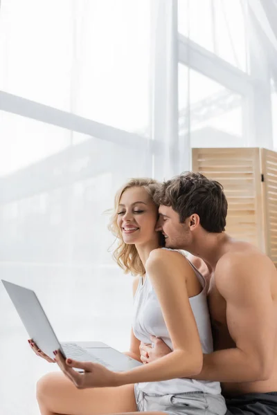 Young muscular man hugging cheerful blonde girlfriend with laptop at home