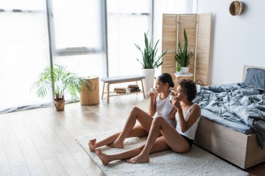 Tam boy Afro-Amerikan lezbiyen çift yatağın yanında oturup kahve içiyorlar.