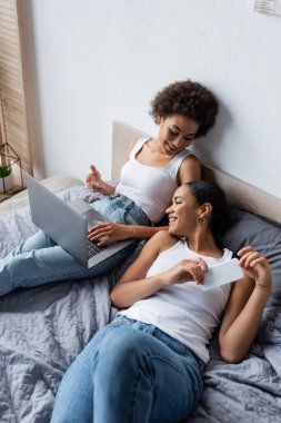 Dizüstü bilgisayarı olan mutlu Afro-Amerikan lezbiyen kadın yatak odasında akıllı telefon tutan kız arkadaşıyla konuşuyor. 
