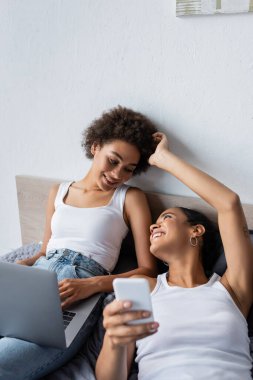 Pozitif Afro-Amerikan lezbiyen kadın akıllı telefon tutuyor ve yatak odasında kız arkadaşının saçını okşuyor.