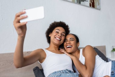 Mutlu Afro-Amerikan lezbiyen çift yatak odasında akıllı telefondan selfie çekiyor.