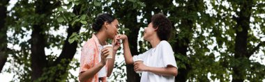 joyful african american lesbian woman holding coffee to go and kissing hand of girlfriend in park, banner clipart