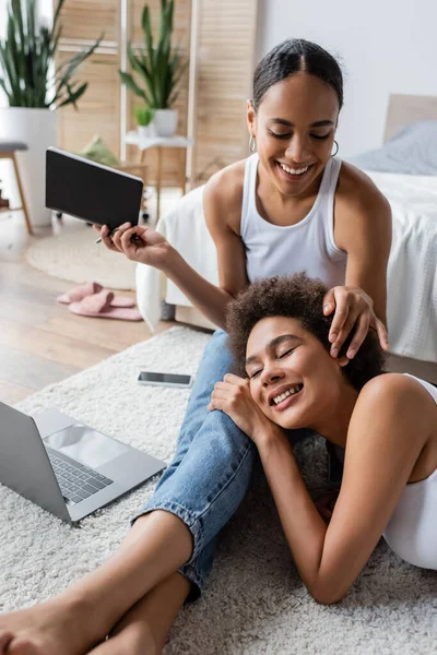 快乐的非洲女同性恋美国女人一边抚摸着女朋友的头发一边拿着笔记本靠近笔记本电脑 — 图库照片