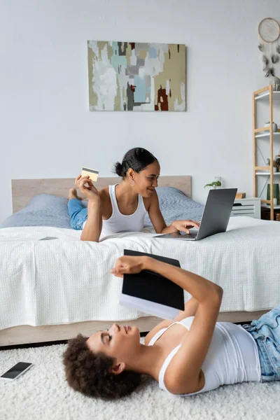アフリカ系アメリカ人の若い女性がラップトップとクレジットカードでオンラインショッピングの近くのカーペットの上に横たわっています — ストック写真