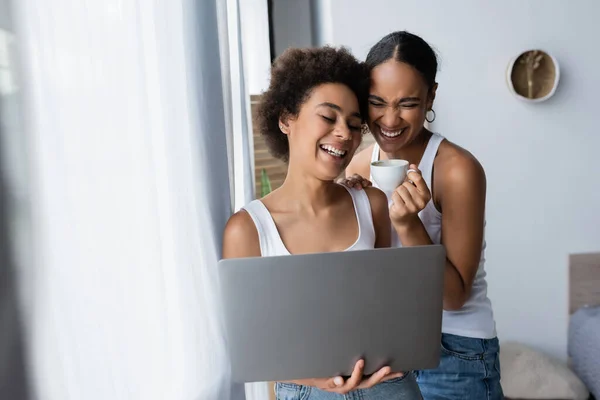 快乐的非洲女同性恋者使用笔记本电脑接近快乐的女朋友 手里拿着咖啡 — 图库照片