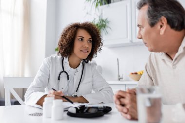 Neşeli Afrikalı Amerikalı doktor orta yaşlı diyabetli bir adamın yanındaki glukometre cihazını işaret ediyor. 