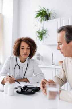 Kıvırcık Afrikalı Amerikalı doktor orta yaşlı hastanın yanındaki glukometreyi işaret ediyor.