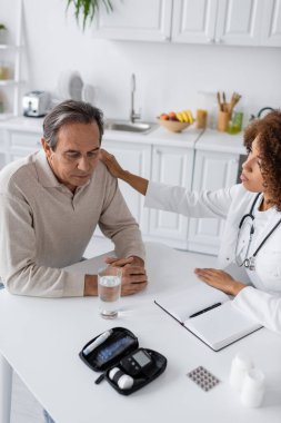 Beyaz önlüklü genç Afrikalı Amerikalı doktor diyabeti olan üzgün orta yaşlı adamı sakinleştiriyor. 