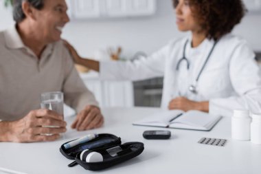 Glikoz ölçer ve tükenmez kalem cihazları, bulanık arka planda diyabeti olan Afrikalı Amerikalı doktor ve orta yaşlı adamın yanında. 