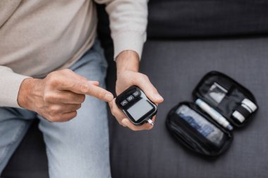 partial view of middle aged man with diabetes pointing at glucose meter device and sitting on sofa  clipart