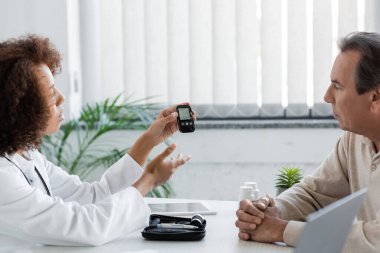 Genç Afrikalı Amerikalı doktor, klinikte diyabeti olan orta yaşlı bir adamın yanındaki glukometreyi işaret ediyor. 