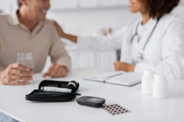 diabetes kit with glucose meter and lancet pen devices near african american doctor and patient on blurred background