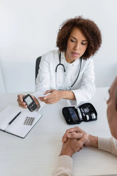 stock image young african american doctor pointing at glucometer near middle aged man with diabetes in clinic 