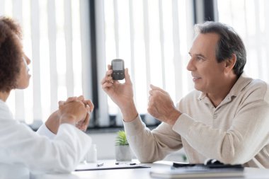 Bulanık Afrikalı Amerikalı bir doktorun kliniğinde glukometre tutan olgun bir hasta var. 