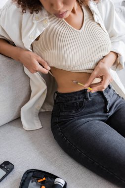 Cropped view of african american woman doing insulin injection near diabetes kit on couch  clipart