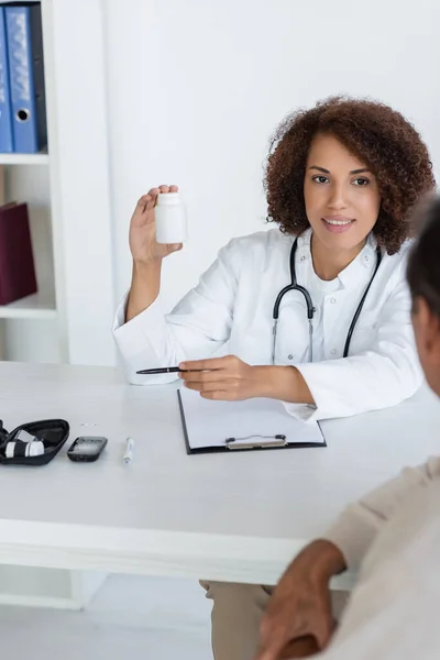 stock image Smiling african american doctor holding pills near blurred patient and diabetes kit in hospital 