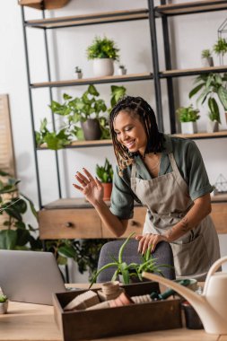 smiling african american florist in apron waving hand near laptop during video call in flower shop clipart