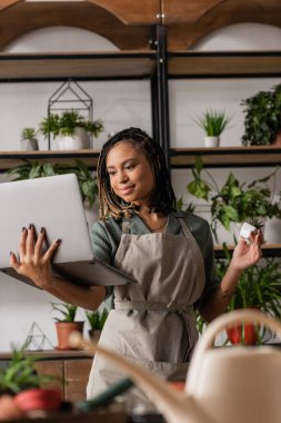 smiling african american woman holding laptop and potted plant during video call near blurred rack with flowerpots clipart