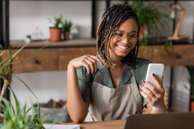 Neşeli Afro-Amerikan çiçekçi, son moda saç stili ve gözlükleriyle çiçekçide cep telefonuna bakıyor.